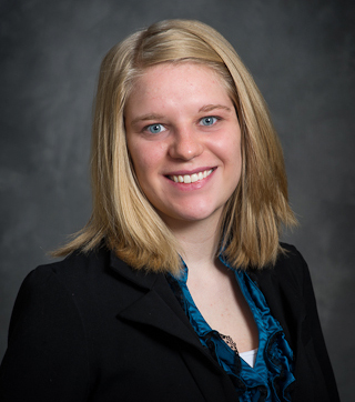 Amanda Huffman, with shoulder-length blonde hair, stands confidently in a black jacket and blue blouse, smiling warmly against a dark, neutral background.